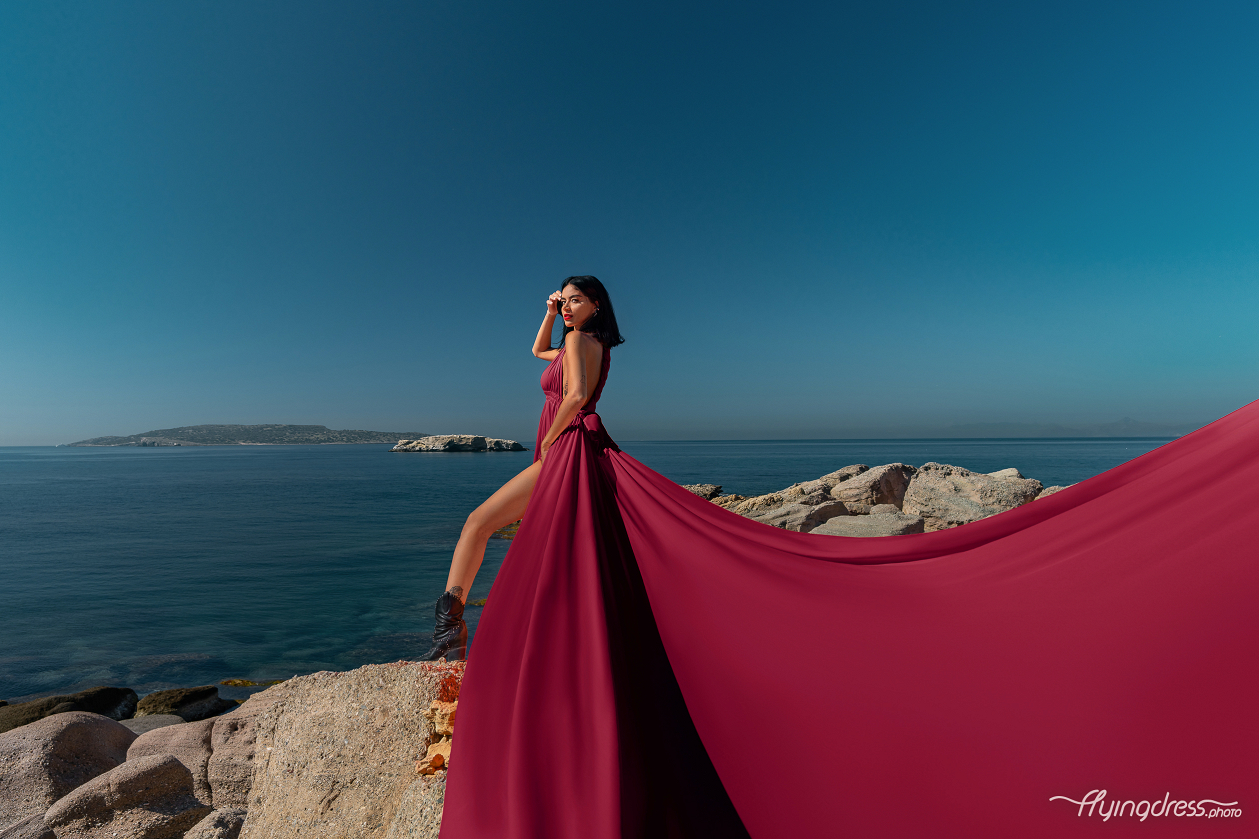 A woman in a flowing burgundy dress and black boots stands poised on rocky cliffs with the expansive blue sea and distant islands of Kavouri, Athens in the background, creating a striking and elegant scene.